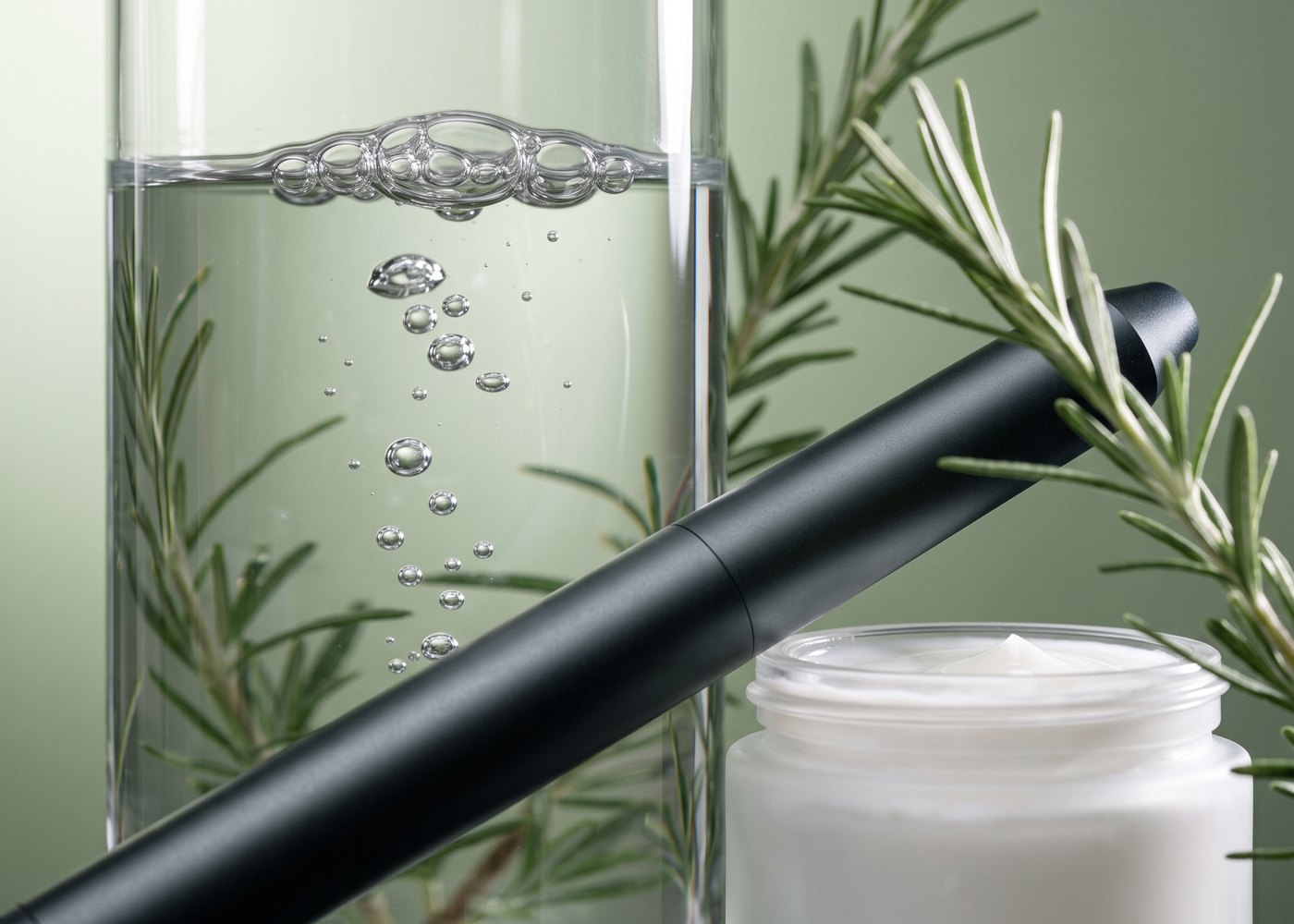 Offener Cremetiegel, ein Glas mit klarem Wasser und aufsteigenden Bläschen und ein dunkles Laser-Handstück auf salbeigrünem Grund, dazwischen Rosmarinzweige