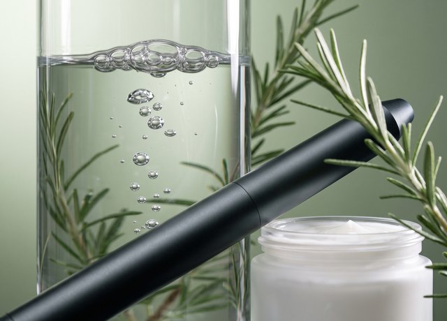 Offener Cremetiegel, ein Glas mit klarem Wasser und aufsteigenden Bläschen und ein dunkles Laser-Handstück auf salbeigrünem Grund, dazwischen Rosmarinzweige