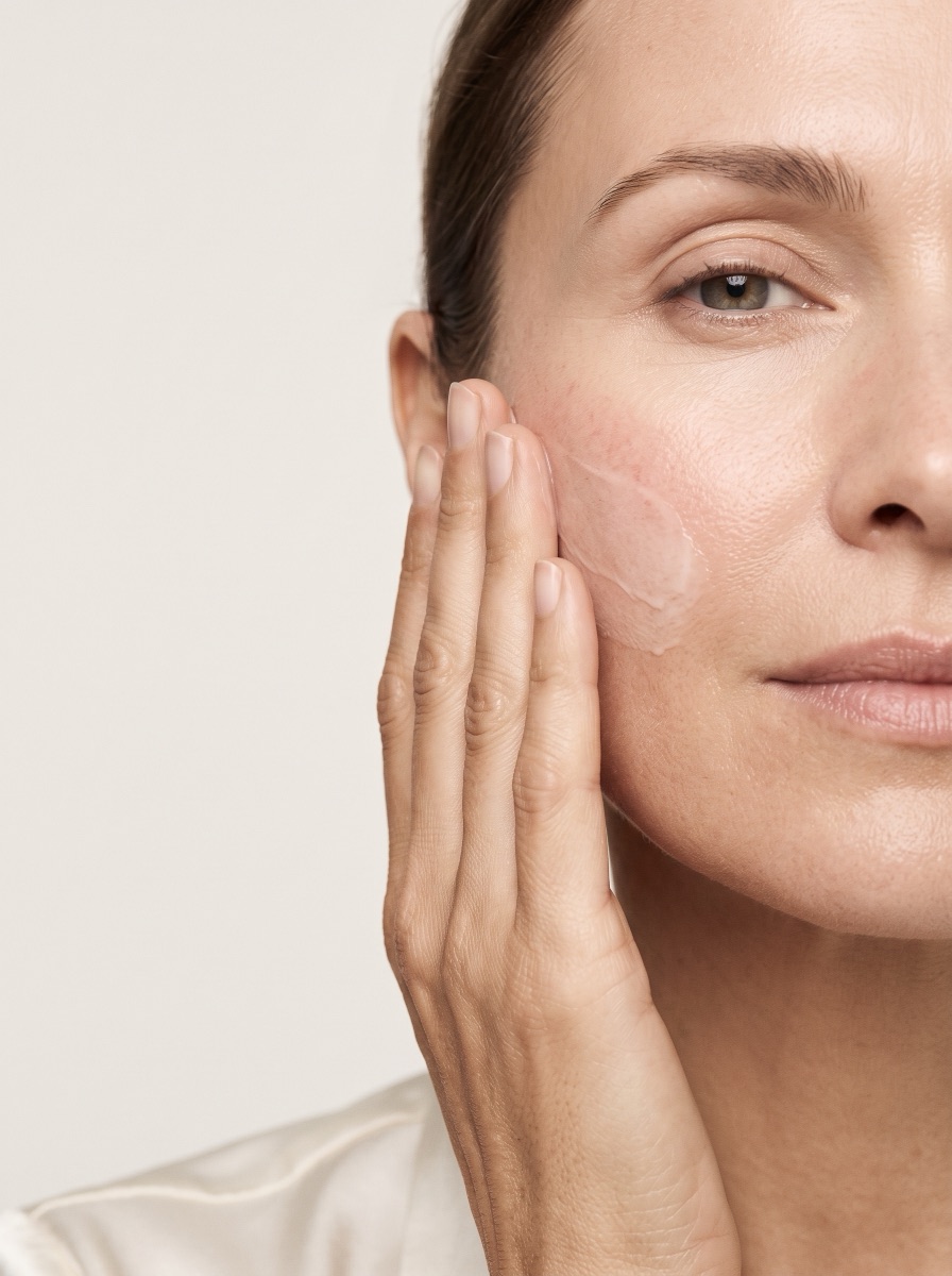Gesicht einer Frau im Anschnitt vor hellem Grund, die Wange leicht gerötet, mit den Fingerspitzen trägt sie eine Creme auf